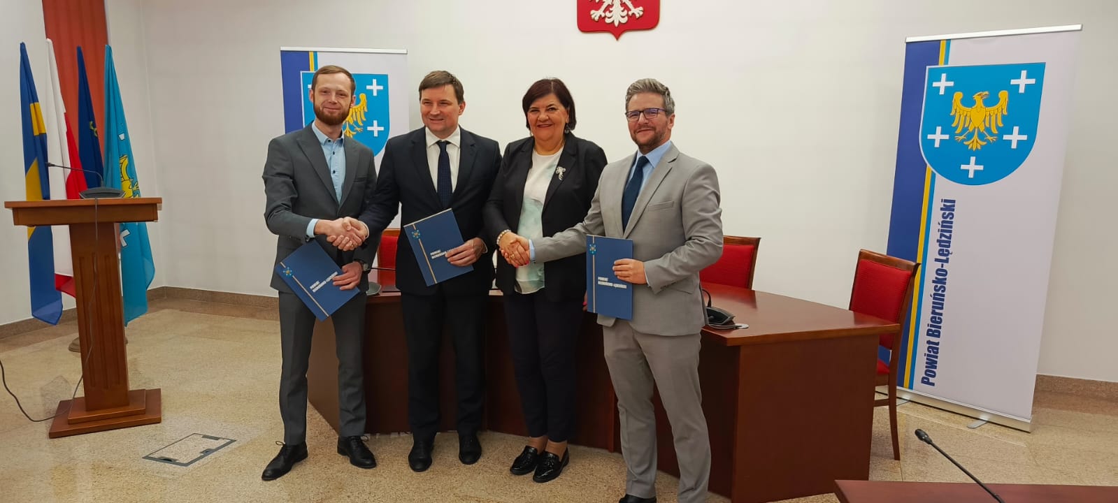 Trzej mężczyźni oraz kobieta stojący przed półokrągłym biurku. Osoby ściskają sobie ręce w geście gratulacji. Na ścianie widoczne godło Polski. Po prawej i lewej stronie stoją pionowe plansze z umieszczonym po lewej stronie herbem powiatu (na niebieskim tle żółty orzeł, wokół pięć białych krzyży). W tle z lewej strony za mównicą stoi stojak z flagami.