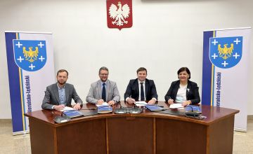 Trzej mężczyźni oraz kobieta siedzący przy półokrągłym biurku podpisujące dokument. Na ścianie widoczne godło Polski. Po prawej i lewej stronie stoją pionowe plansze z umieszczonym po lewej stronie herbem powiatu (na niebieskim tle żółty orzeł, wokół pięć białych krzyży)