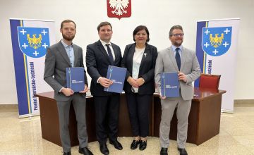 Trzej mężczyźni oraz kobieta stojący przed półokrągłym biurku. Panowie trzymają w rękach dokument. Na ścianie widoczne godło Polski. Po prawej i lewej stronie stoją pionowe plansze z umieszczonym po lewej stronie herbem powiatu (na niebieskim tle żółty orzeł, wokół pięć białych krzyży)