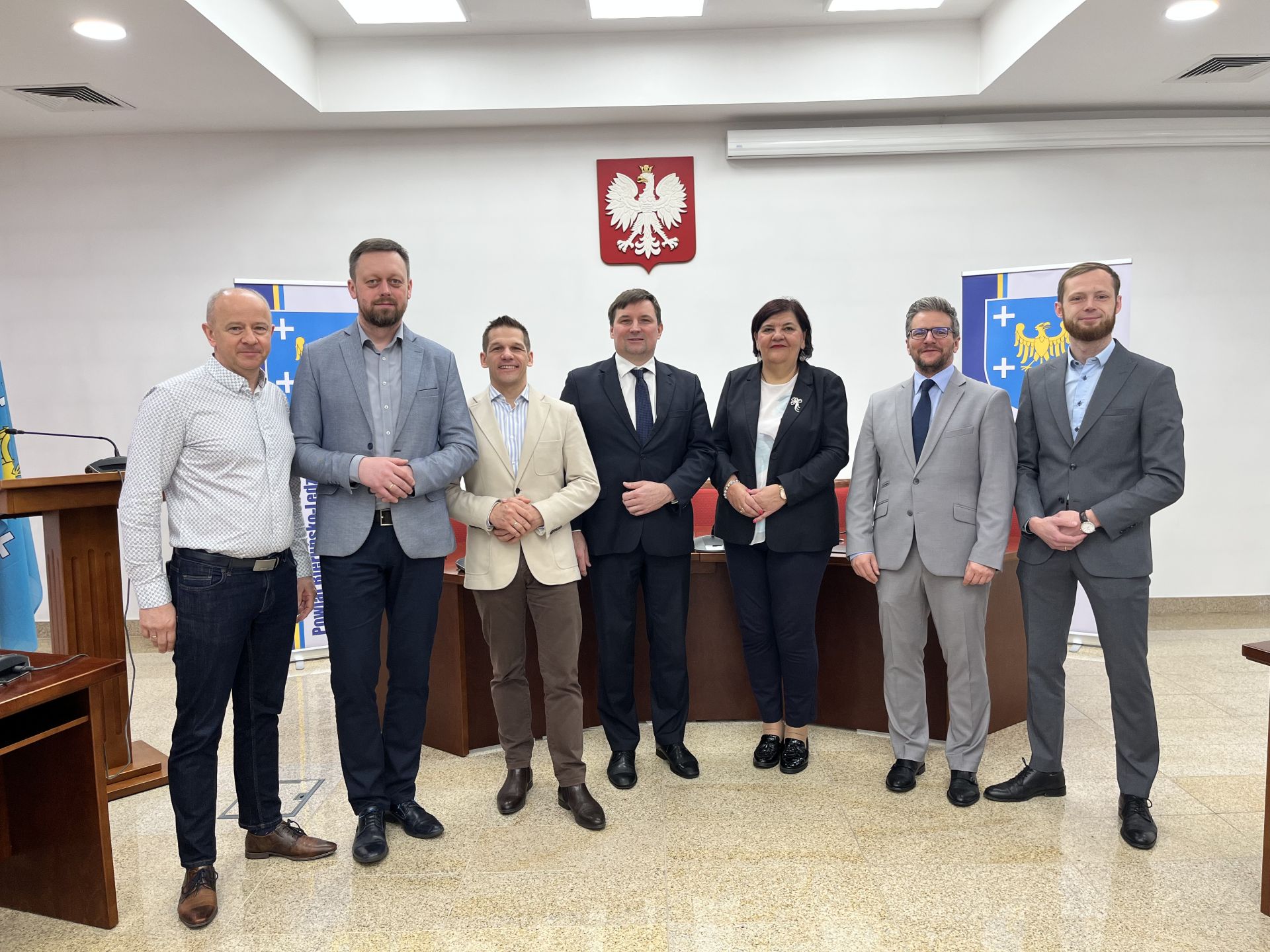 Zdjęcie wykonane na sali konferencyjnej. Przed półokrągłym stołem stoi siedem osób. Sześciu mężczyzn oraz kobieta. Wszyscy są uśmiechnięci i elegancko ubrani. W tle na ścianie widać godło Polski. Po prawej i lewej stronie widoczne są częściowo plansze przedstawiające herb powiatu. Jest to żółty orzeł wokół niego pięć białych krzyży. Wszystko na niebieskim tle.