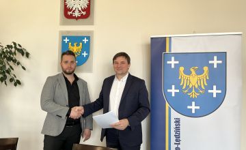 Dwóch mężczyzn stojących na tle planszy z herbem powiatu. Panowie ściskają sobie prawe ręce w geście gratulacji. Mężczyzna stojący z prawej strony trzyma w ręce dokument.
