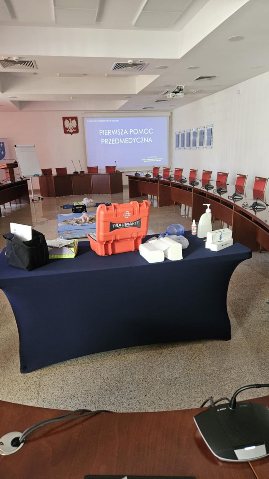 Sala konferencyjna, pod ścianami ułozone półokrągłe stoły, na ścianie wyświetlany jest napis pierwsza pomoc przedmedyczna. Na pierwszym planie widać stół z niebieskim pokrowcem. Na stole leży apteczka, defibrylator, płyn do dezynfekcji. Na ziemi częściowo widać rozłożony koc z fantomami.