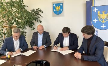 Czterech mężczyzn elegancko ubranych siedzących przy stole. Panowie podpisują dokumenty. Z lewej strony częściowo widać duży kwiat z drobnymi listkami. Na ścianie wisi godło Polski oraz herb powiatu. Z prawej strony plansza z herbem powiatu.