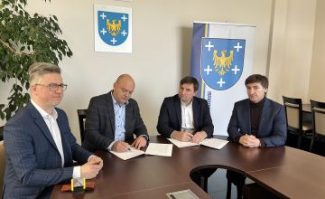 Czterech mężczyzn elegancko ubranych siedzących przy stole. Panowie podpisują dokumenty. Z lewej strony częściowo widać duży kwiat z drobnymi listkami. Na ścianie wisi godło Polski oraz herb powiatu. Z prawej strony plansza z herbem powiatu.