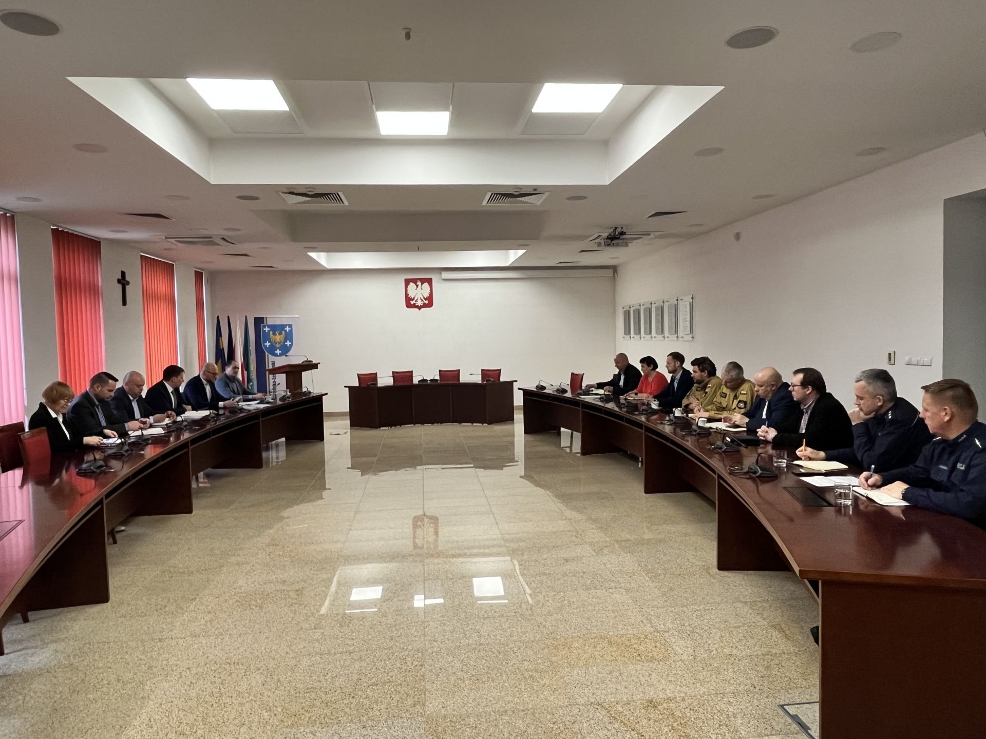 Sala konderencyjna. przy półokragłych stołach znajdujących się po prawej i lewej stronie siedzą ludzie. Z przodu sali znajduje się stól z czterema krzesłami. Z lewej strony stoi mównica. Obok mównicy znajduje się stojak z flagami. Na ścianie wisi godło Polski. Z prawej strony na ścianie widać wiszące tablice. Po przeciwnej stronie znajdują się okna z roletami. Na ścianie wisi krzyż. 