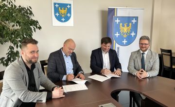 Czterech mężczyzn elegancko ubranych siedzących przy stole. Panowie podpisują dokumenty. Z lewej strony częściowo widać duży kwiat z drobnymi listkami. Na ścianie wisi godło Polski oraz herb powiatu. Z prawej strony plansza z herbem powiatu. 