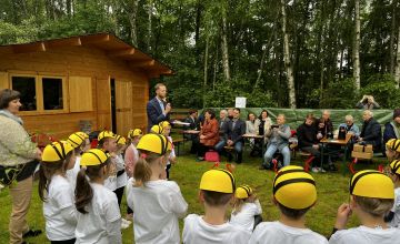 Występ dzieci ubranych w pszczele czapeczki. W tle drewniany budynek, zielone drzewa. Przy czterech stołach siedzą pszczelarze oraz pozostali goście.