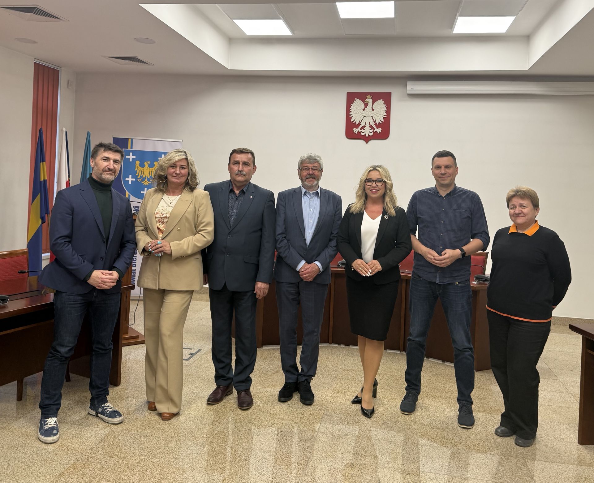 Zdjęcie przedstawiające salę konferencyjną. Na pierwszym planie w rzędzie stoi 7 osób. Na białej ścianie za nimi wisi godło Polski. Z lewej strony widać fragment ścianki z herbem (na niebieskim tle żółty orzeł a wokół niego pięć białych krzyży). Po bokach oraz za stojącymi ludźmi częściowo widoczne są stoły. 