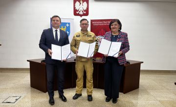 sala konferencyjna. Na ścianie wisi godło Polski. Przed półokrągłym stołem stoją trzy osoby trzymające dokumenty. Za nimi stoją banery. Ze lewej strony baner z herbem powiatu, z prawej ścianka straży pożarnej. 
