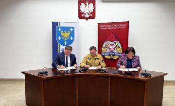 sala konferencyjna. Na ścianie wisi godło Polski. Przy półokrągłym stole siedzą trzy osoby podpisujące dokumenty. Za nimi stoją banery. Ze lewej strony baner z herbem powiatu, z prawej ścianka straży pożarnej. 
