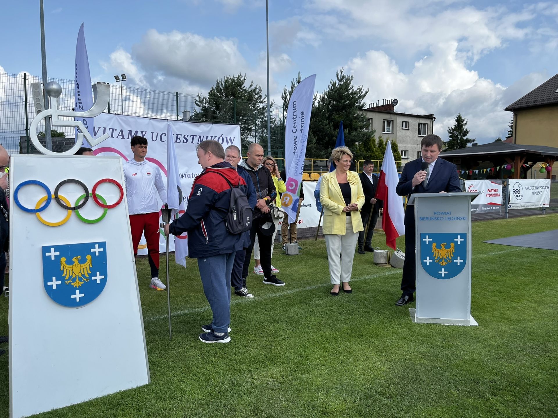 Zdjęcie przedstawiające postument z kołami olimpijskimi. Na postumencie znicz olimpijski w kształcie osoby na wózku inwalidzkim. Przy mównicy przemawia osoba.