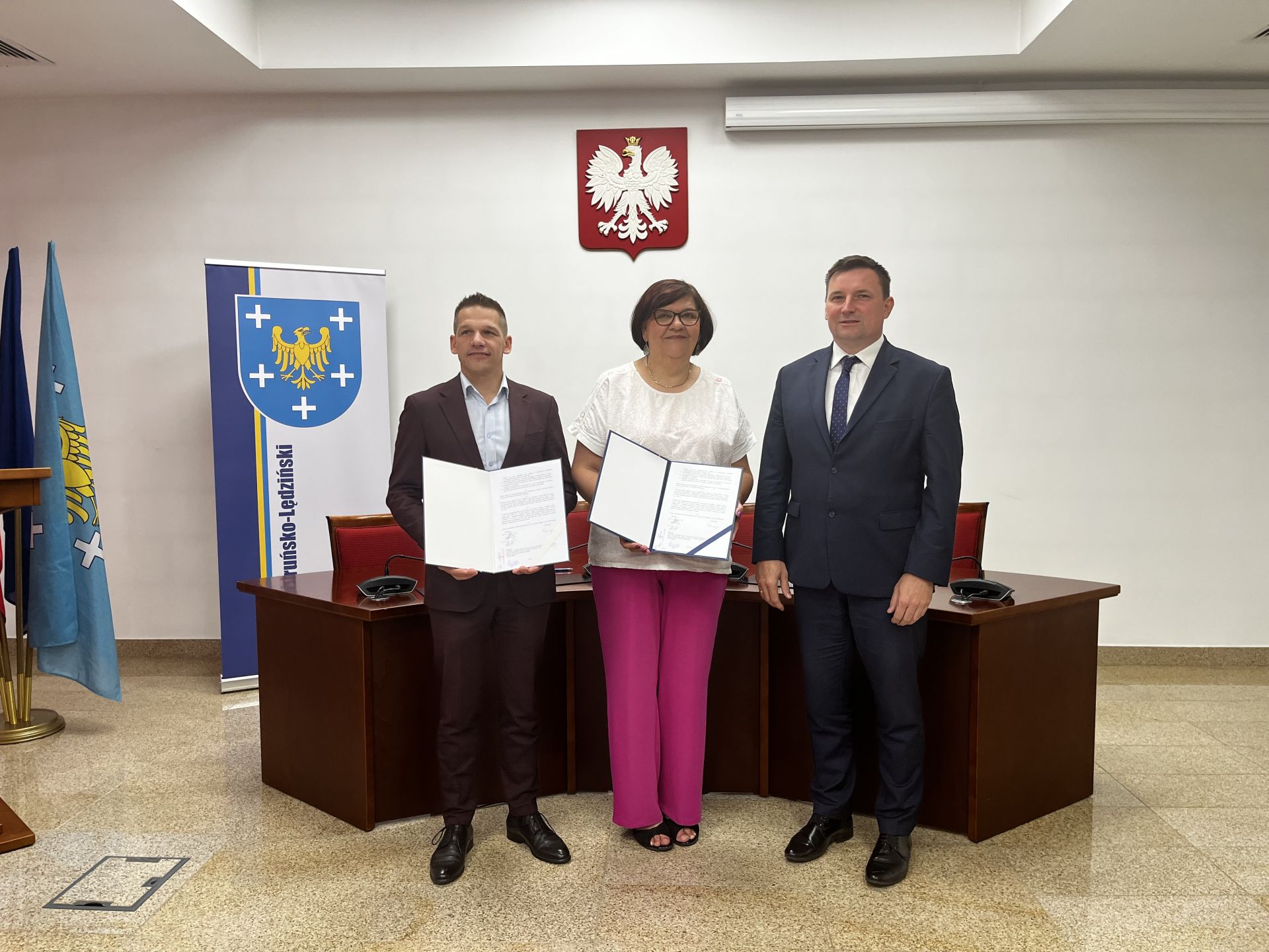 Sala konferencja. Przed półokrągłym stołem stoją trzy osoby. Dwie z nich trzymają w rękach otwarte teczki z dokumentami. W tle na ścianie wisi godło. Z lewej strony ustawione są flagi oraz ścianka z herbem powiatu.