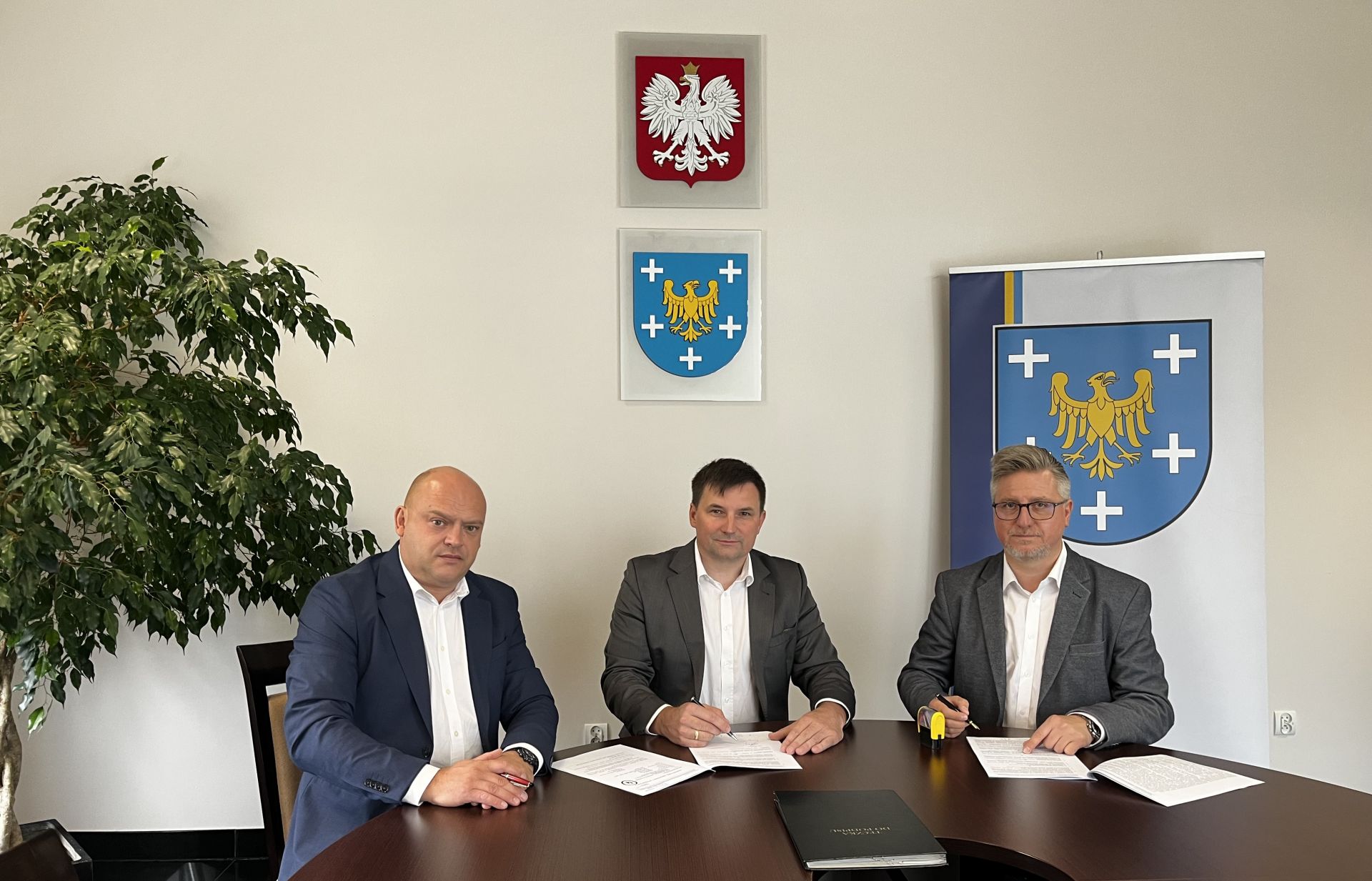 Trzy osoby siedzące przy stole podpisują dokumenty. Na ścianie wisi herb powiatu oraz godło.