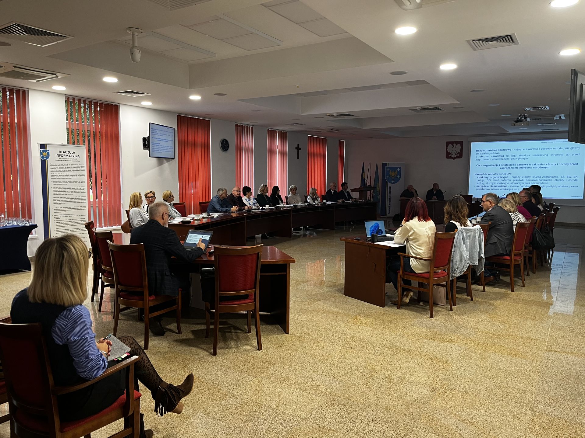 Zdjęcie przedstawiające grupę osób siedzących przy dwóch stołach. Na ekranie wyświetlany jest tekst.
